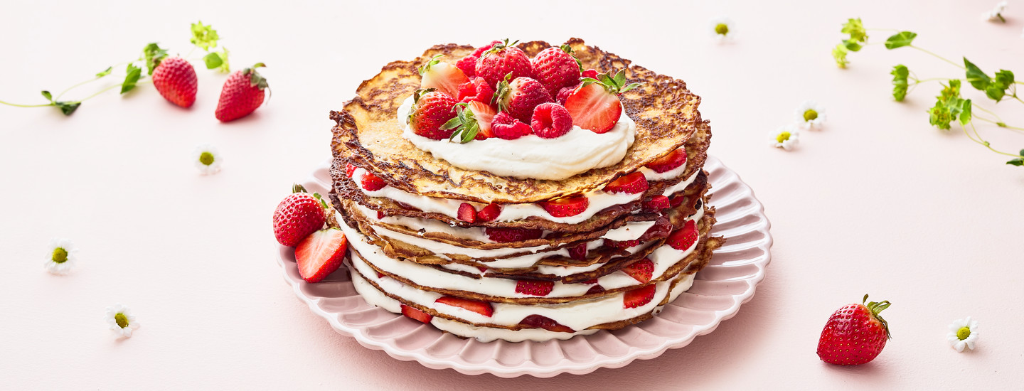 Lettukakku vaaleanpunaisella lautasella. Lautasella on pino lettuja, joiden väliin on lisätty kermavaahtoa ja tuoreita mansikoita. K-Ruoka jälkiruokaresepti.