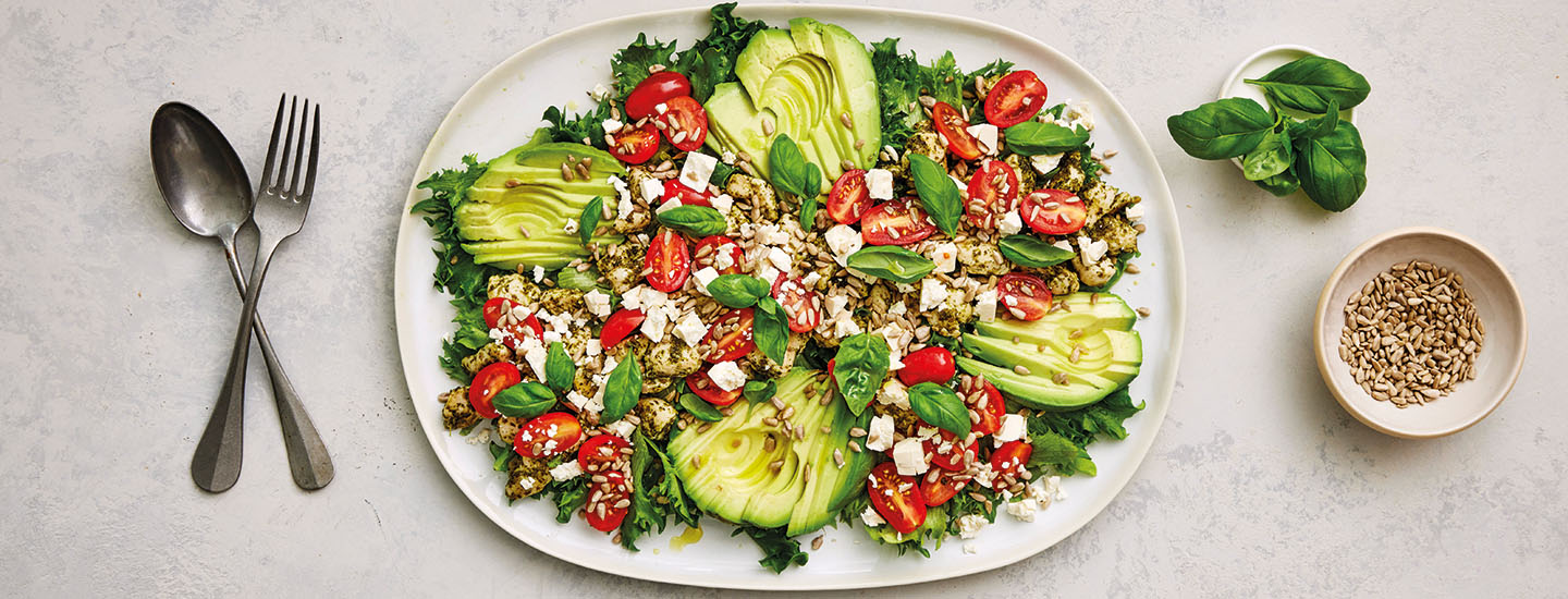 Kana-avokadosalaattia laakealla valkoisella lautasella. Avokadot on siivutettu ja ne on aseteltu neljään eri sektoriin lautaselle. Salaatin joukossa on jääsalaattia, pestokanaa, miniluumutomaatteja ja salaattijuustoa. K-Ruoka salaattiresepti.