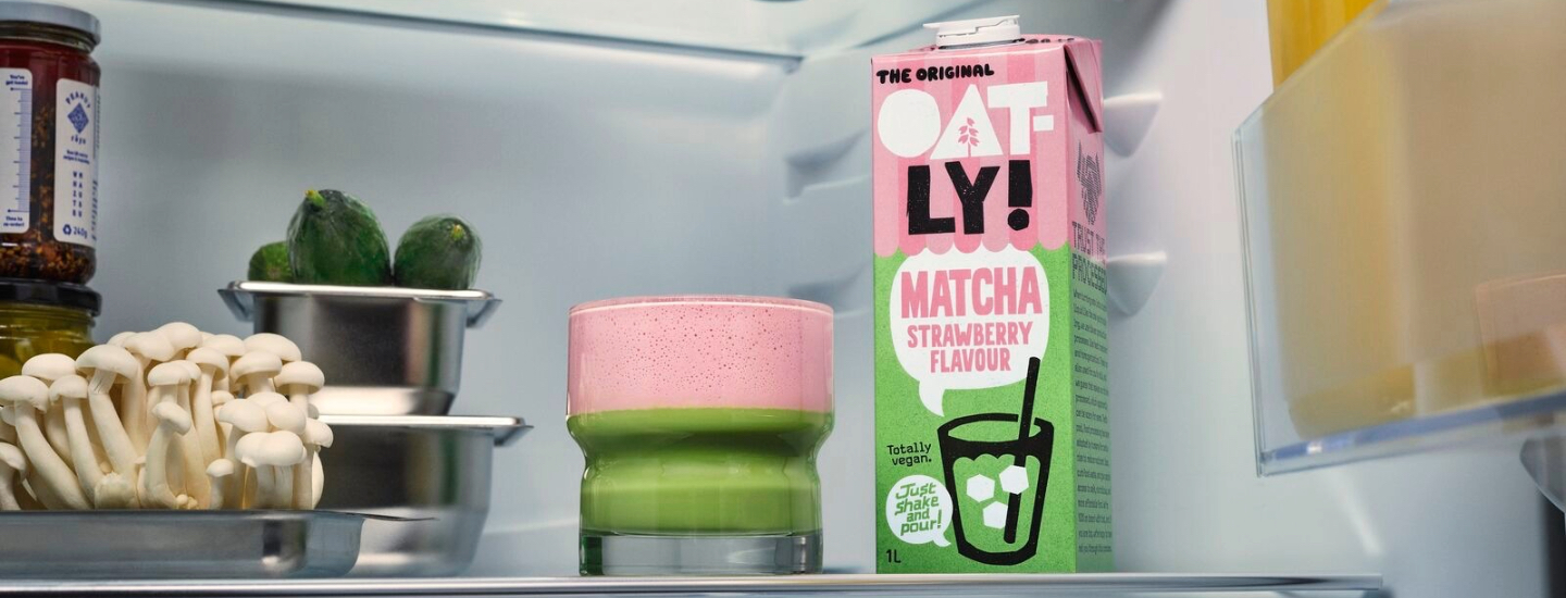 Jääkaapissa säilykepurkkeja, vihanneksia ja etualalla Oatly Matcha Strawberry flavour purkki ja lasi, jossa pohjalla vihreää ja päällä vaaleanpunaista juomaa.