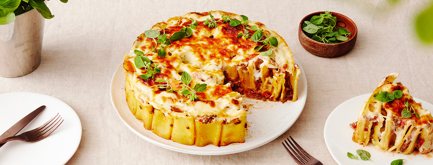 Putkipastalasagnea valkoisella pyöreällä tarjoilulautasella. Lasagnen päälle on lisätty valkokastike- ja juustokerros. Lasagne on koristeltu tuoreilla yrteillä. K-Ruoka arkiruokaresepti.