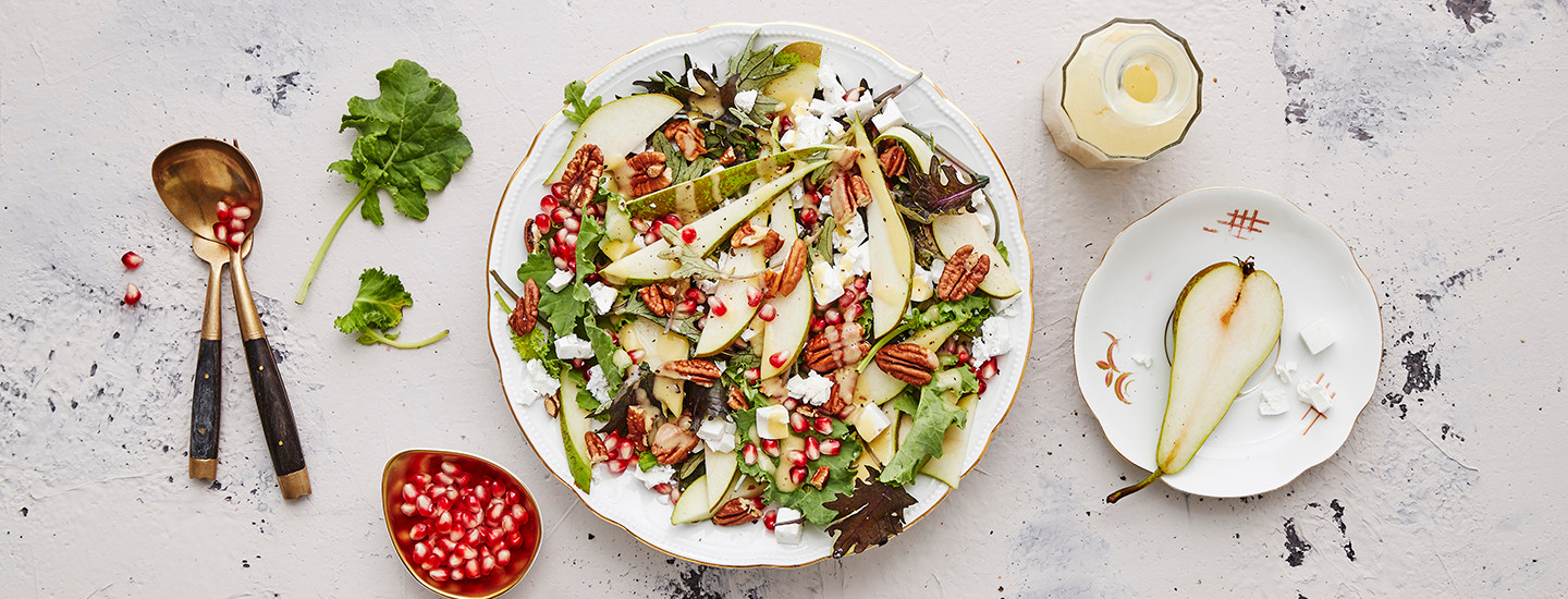Päärynä-lehtikaalisalaatti valkoisella lautasella. Salaatissa on lisäksi päärynäsiivuja ja vuohenjuustomurua. Lautasen vieressä on pieni kulho granaattiomenan siemeniä sekä salaatinottimet. K-Ruoka salaattiresepti.