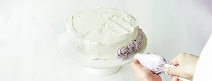 Täytekakku, jonka päälle on levitetty kermavaahtoa. Kakun sivuun ollaan tekemässä vaaleanliiloja ruusupursotuksia.