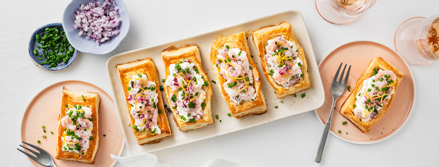 Skagenpiirakoita valkoisella tarjoilulautasella. Skagenpiirakat on pienessä koossa ja suorakulmion muotoisia. Piirakat on täytetty katkarapusalaatilla, katkaravuilla, punasipulilla, raastetulla sitruunankuorella ja ruohosipulilla. K-Ruoka piirakkaresepti.