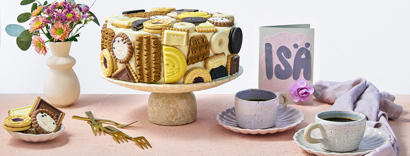 Keksikakku vaalealla kakkutarjottimella. Kakkuun on aseteltu päälle erilaisia keksejä. Kakun vieressä on isänpäiväkortti, jossa lukee "ISÄ", sekä kaksi vaaleansinistä kahvikuppia. K-Ruoka isänpäivän kakkuresepti.