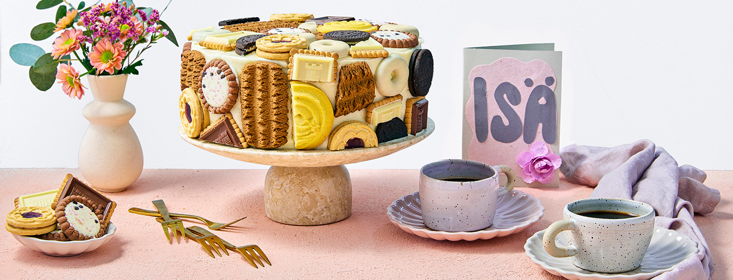 Keksikakku vaalealla kakkutarjottimella. Kakkuun on aseteltu päälle erilaisia keksejä. Kakun vieressä on isänpäiväkortti, jossa lukee "ISÄ", sekä kaksi vaaleansinistä kahvikuppia. K-Ruoka isänpäivän kakkuresepti.