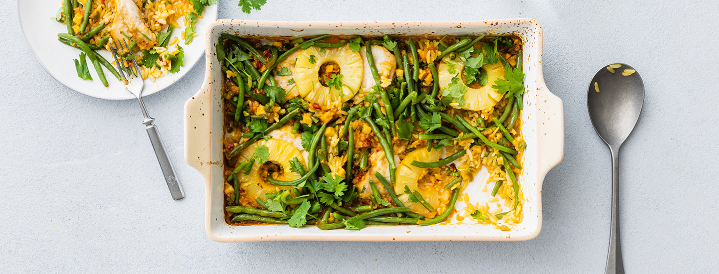 Havaijin kanavuokaa valkoisessa uunivuoassa. Vuokaruoassa on kanaa, vihreitä papuja, riisiä ja ananasviipaleita. Uunivuoan oikealla puolella on lusikka, vasemmalla puolella lautanen, jossa on kanavuokaa. K-Ruoka arkiruokaresepti.