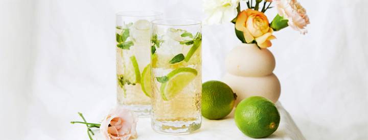 Kaksi simadrinkkiä vaalealla liinalla peitetyllä pöydällä kahden limen ja kukkien kanssa.