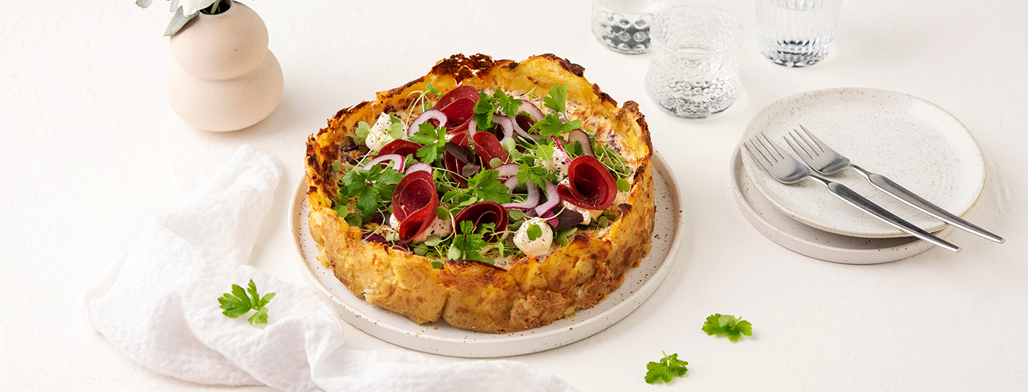 Lyttyperunapiirakka valkoisella pyöreällä tarjoilulautasella. Piirakan pohja on tehty lytätyistä perunoista ja pohjan reuna nousee korkeammalle kuin piirakan täyte. Piirakkan päälle on aseteltu kylmäsavuporoviipaleita ja tuoreita yrttejä. K-Ruoka piirakkaresepti.