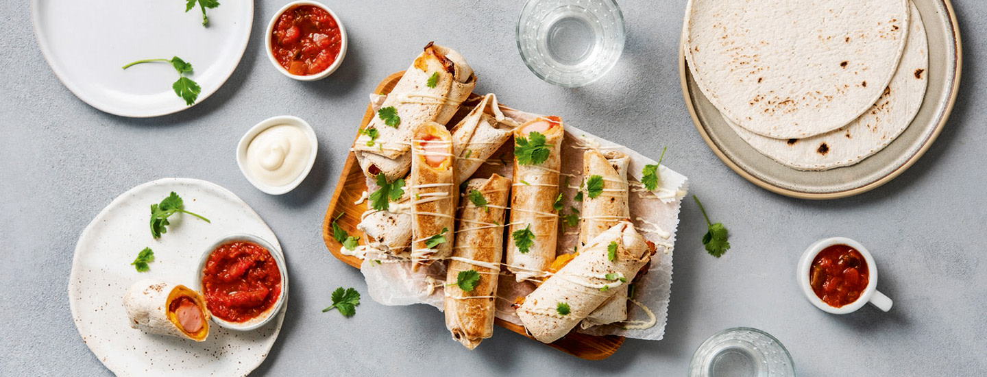 Tortillaan tehtyjä hot dogeja puisella tarjoiluastialla. Kuvassa myös lautanen, jossa on tortillalettuja sekä pieniä salsakastikekuppeja. K-Ruoka arkiresepti.