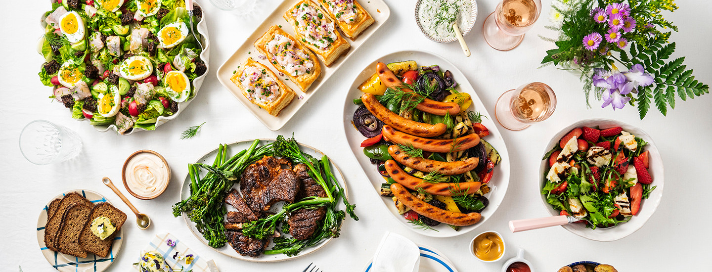 Katettu pöytä, jolla on erilaisia ruokia tarjoiluastioissa. Isossa valkoisessa kulhossa on juhannussalaattia, jossa on vihersalaattia, keitettyjä kananmunia, silliä ja tomaatteja. Suorakulmion muotoisella tarjoiluastialla on skågenpiirakoita. Laakealla valkoisella tarjoiluastialla on grillattuja makkaroita kasvispedillä. Toisella valkoisella laakealla tarjoiluastialla on grillattuja kasslerpihvejä ja varsiparsakaaleja. Pienemmässä valkoisessa kulhossa on salaattia, jossa on vesimelonia, halloumia ja vihersalaattia. K-Ruoka juhannustarjoilut.