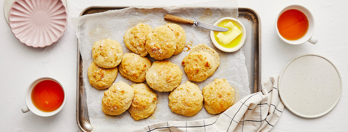 Gluteenittomia sämpylöitä uunipellin päällä. Sämpylöiden vieressä on pieni astia, jossa on voita hieman sulaneena. K-Ruoka gluteeniton resepti.