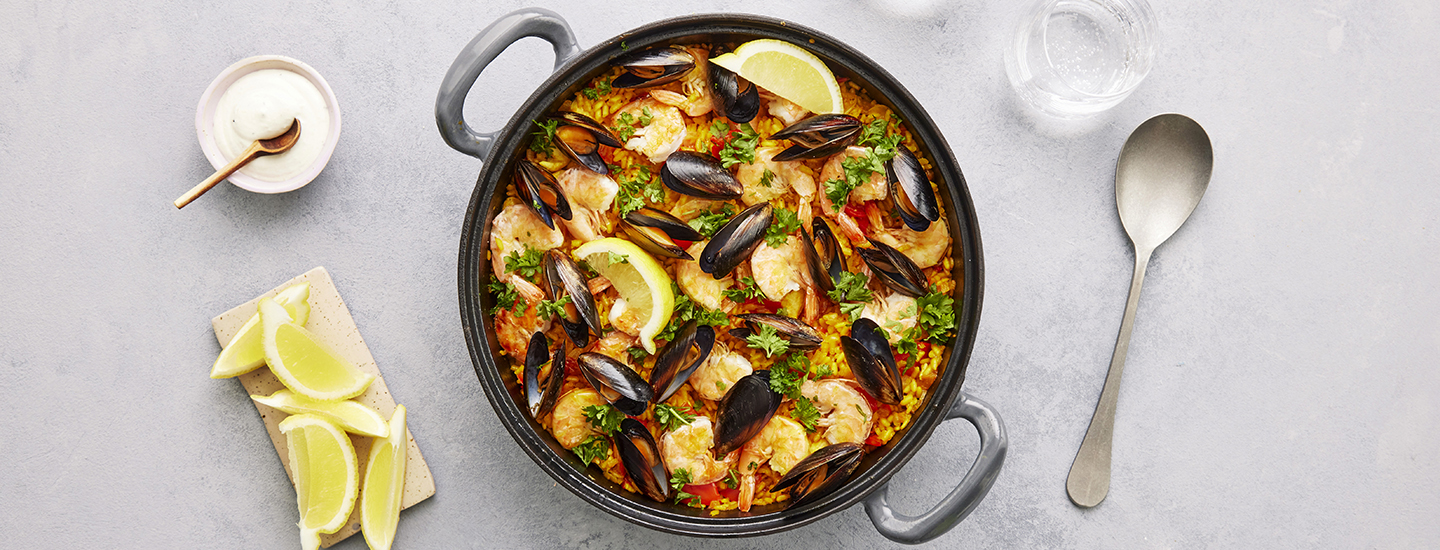 Paellaa valurautakattilassa. Paellan pinnalla on simpukoita, katkarapuja, tuoreita yrttejä ja sitruunalohkoja. K-Ruoka resepti.