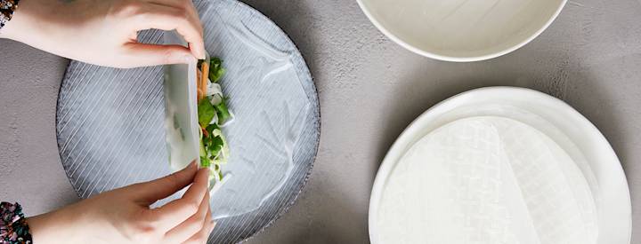 Sinisellä lautasella on kasteltu riisipaperi, jonka keskelle on aseteltu erilaisia täytteitä, kädet ovat taittelemassa riisipaperia täytteiden päälle.