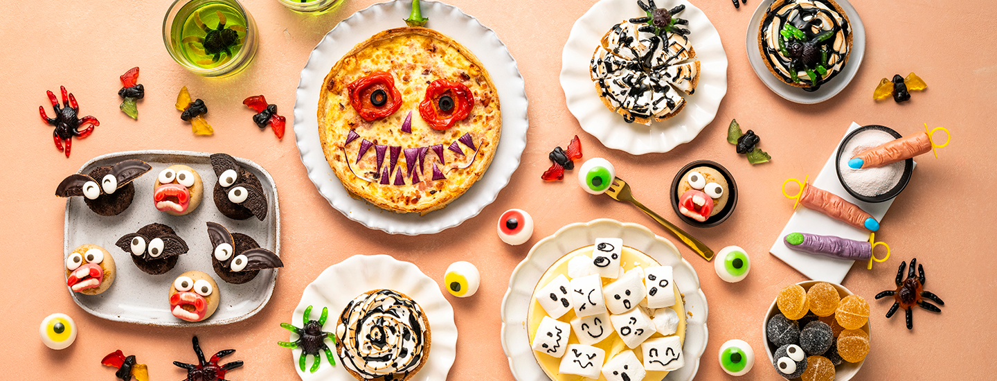 Erilaisia halloween-herkkuja tarjoilulautasilla. Muffinsseja, joissa on puolikkaat keksit lepakonsiipinä. Keksejä, joilla on kasvot. Suolainen piirakka, jolle on lisätty silmät ja terävät hampaat kasviksista.