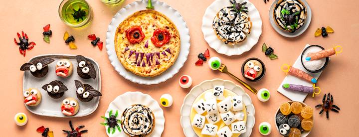 Erilaisia halloween-herkkuja tarjoilulautasilla. Muffinsseja, joissa on puolikkaat keksit lepakonsiipinä. Keksejä, joilla on kasvot. Suolainen piirakka, jolle on lisätty silmät ja terävät hampaat kasviksista.