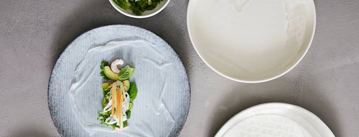 Sinisellä lautasella on kasteltu riisipaperi, jonka keskelle on aseteltu erilaisia täytteitä, riisipaperista ja täytteistä taitellaan vietnamilainen kesärulla.