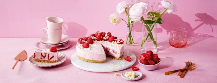 Vadelmainen juustokakku valkoisella kakkutarjottimella. Juustokakun täytteeseen on vadelmanpuolikkaita. Juustokakku on koristeltu valkosuklaalla ja tuoreilla vadelmilla. K-Ruoka juustokakkuresepti.