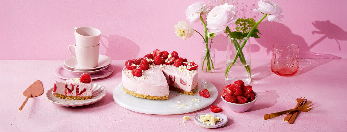 Vadelmainen juustokakku valkoisella kakkutarjottimella. Juustokakun täytteeseen on vadelmanpuolikkaita. Juustokakku on koristeltu valkosuklaalla ja tuoreilla vadelmilla. K-Ruoka juustokakkuresepti.