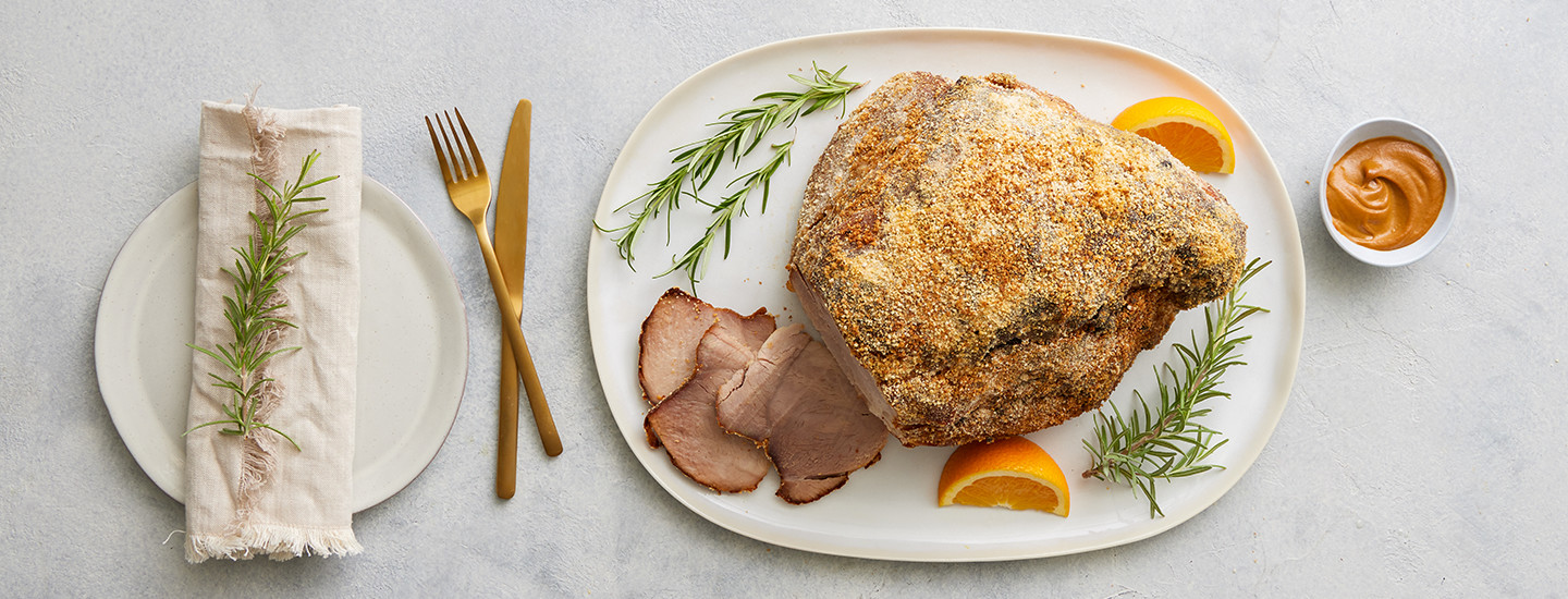 Joulukinkku valkoisella tarjottimella. Kinkku on kuorrutettu sinapilla ja korppujauholla. Kinkusta on leikattu kolme siivua, jotka ovat kinkun vierellä tarjottimella. Tarjottimella on myös kaksi appelsiinisiivua sekä kolme rosmariininoksaa. K-Ruoka jouluresepti.