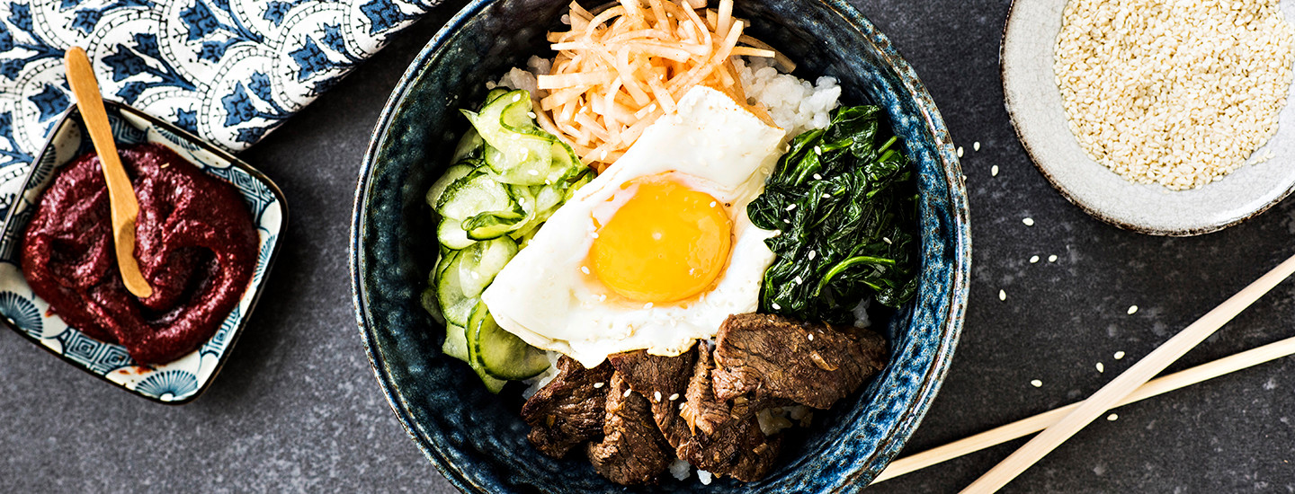 Korealainen bibimbap-annos sinisessä kulhossa. Kulhossa on riisiä, merilevää, kurkkua, lihaa, retiisiä ja paistettu kananmuna keskellä. Vasemmalla puolella kulhoa on pienempi kuppi, jossa on punaista gochujang-chilitahnaa. K-Ruoka korealainen ruokaresepti.