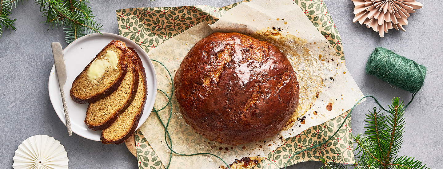 Kokonainen joululimppu leivinpaperin ja lahjapaperin päällä. Joululimpun vieressä on valkoinen lautanen, jossa on kolme siivua limpusta päällekkäin, joista päällimmäinen on voideltu. K-Ruoka jouluresepti.