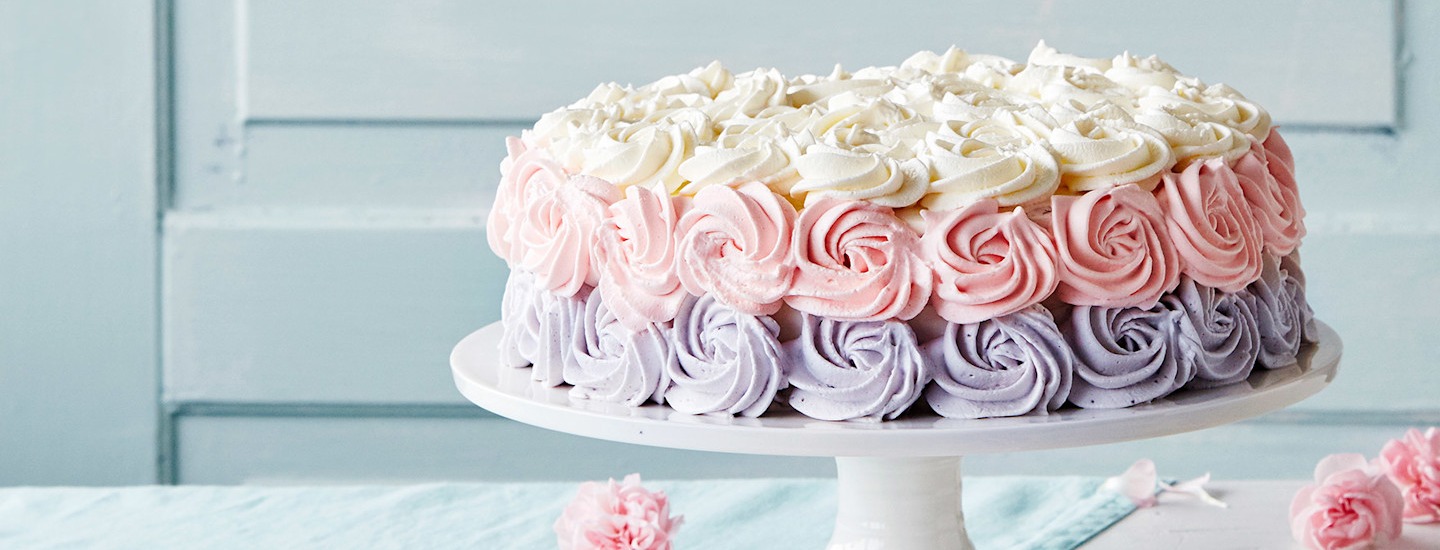 Täytekakku, joka on koristeltu ruusupursotuksilla. Ruusupursotukset ovat valkoisia, vaaleanpunaisia ja vaaleanliiloja. Täytekakku on valkoisen kakkulautasen päällä. K-Ruoka täytekakku.