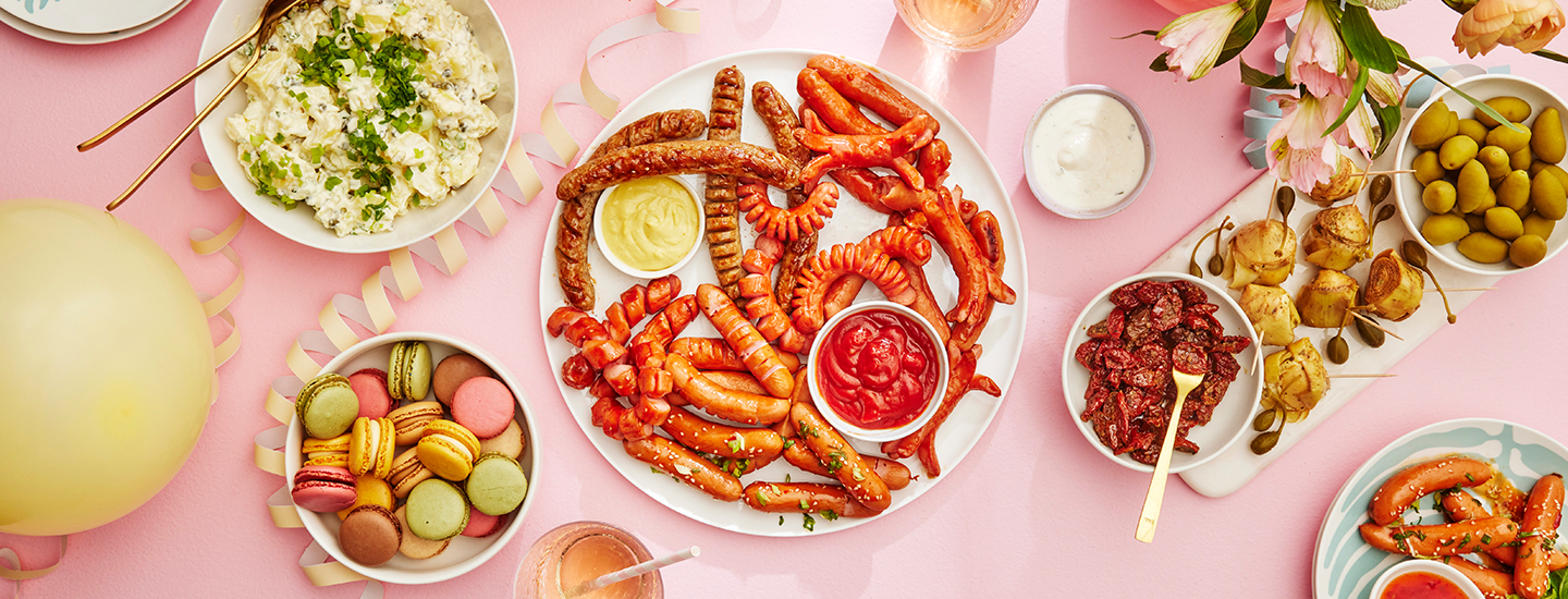 Nakkeja, perunasalaattia, macaroneja ja tapaksia tarjoiltuna pöydällä. Tarjoilujen välissä on erilaisia vappukoristeita. K-Ruoka vappu.