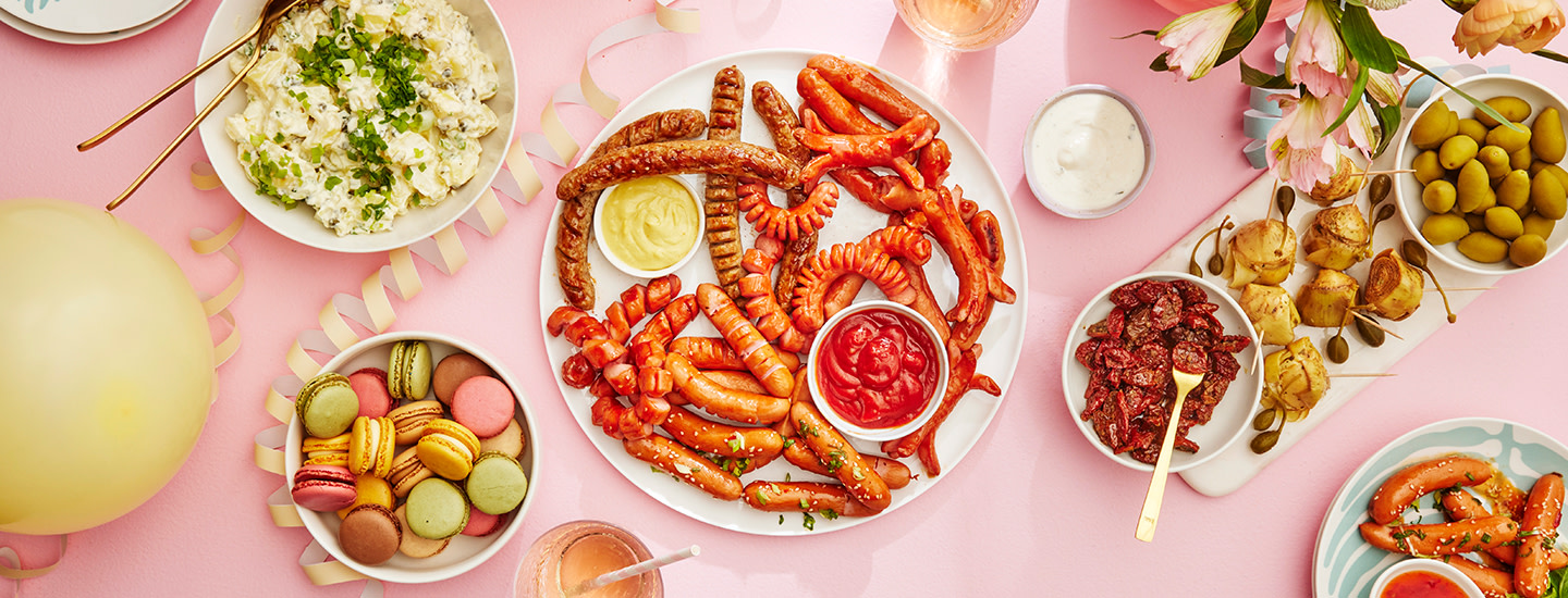 Nakkeja, perunasalaattia, macaroneja ja tapaksia tarjoiltuna pöydällä. Tarjoilujen välissä on erilaisia vappukoristeita. K-Ruoka vappu.