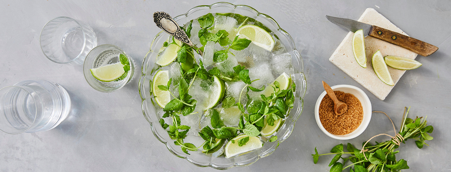 Läpinäkyvää juotavaa läpinäkyvässä lasikulhossa, jossa on seassa jääpaloja, mintunlehtiä ja limeviipaleita.