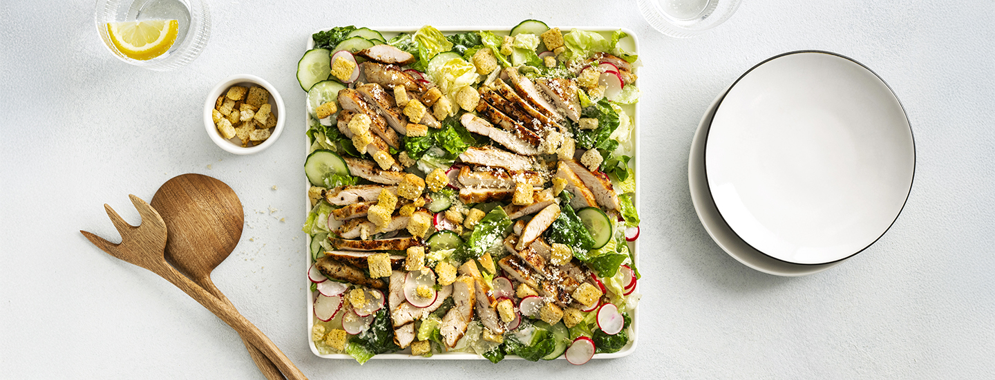 Kanacaesarsalaattia neliönmuotoisella tarjoilulautasella. Kana on grillattu ja viipaloitu. Vihersalaatin, kurkku- ja retiisiviipaleiden ja grillatun kanan lisäksi salaatissa on krutonkeja ja raastettua parmesaania. K-Ruoka salaattiresepti.