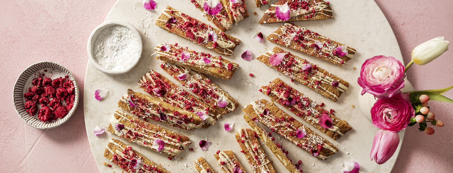 Viipalekeksejä valkoisella pyöreällä tarjottimella. Keksit on koristeltu valkosuklaalla, kuivatuilla vadelmilla ja syötävien kukkien terälehdillä. K-Ruoka keksiresepti.