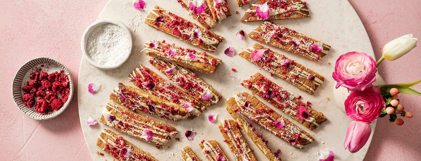 Viipalekeksejä valkoisella pyöreällä tarjottimella. Keksit on koristeltu valkosuklaalla, kuivatuilla vadelmilla ja syötävien kukkien terälehdillä. K-Ruoka keksiresepti.