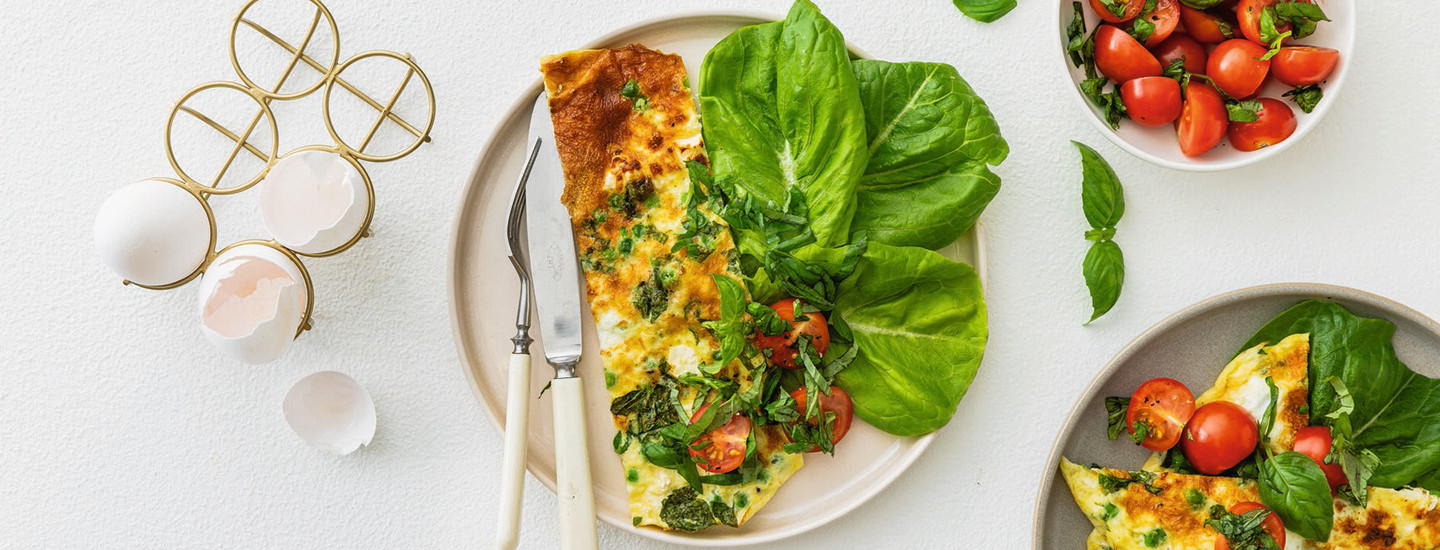 Airfryerissa valmistettua munakasta vaalealla pyöreällä lautasella. Munakkaan vieressä vasemmalla puolella on haarukka ja veitsi. Oikealle puolelle lautasta on laitettu salaatinlehtiä. Munakkaan päälle on lisätty tuoreita yrttejä ja kirsikkatomaatin puolikkaita. K-Ruoka arkiruokaresepti.