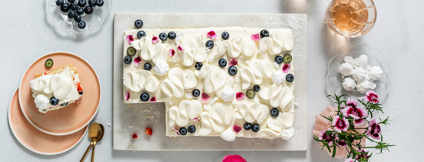 Levykakku vaalealla tarjottimella. Levykakun päälle on lisätty tuorejuusto-kermavaahtoa. Kermavaahdolla on myös tehty pursotukset kakun päälle. Kakku on koristeltu mustikoilla ja neilikan terälehdillä. K-Ruoka kakkuresepti.