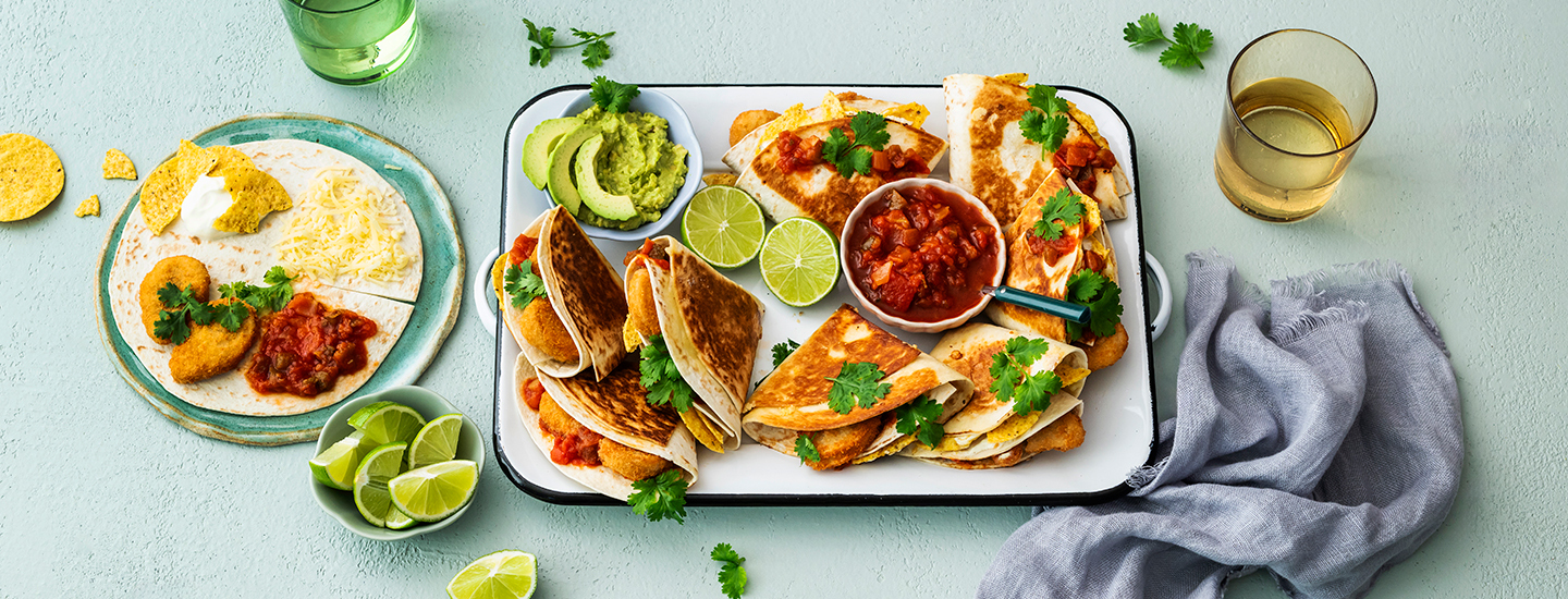 Taiteltuja tortillataskuja tarjottimella. Tortillataskujen sisällä on juustoa, salsaa, kananugetteja ja korianteria. Tarjottimella on tortillataskujen lisäksi pienessä astiassa salsaa ja toisessa pienessä astiassa guacamolea. K-Ruoka texmex-resepti.