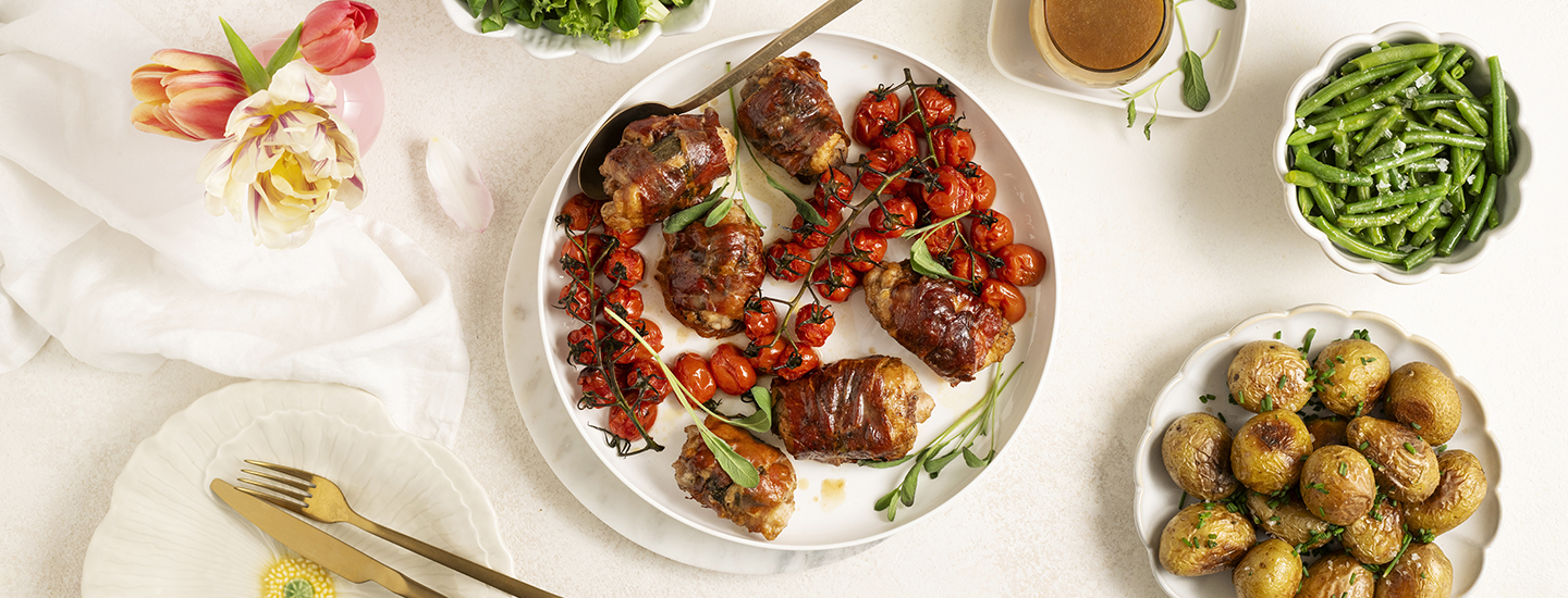 Kanasaltimboccaa, eli kanan paistileikkeitä käärittynä parmankinkkuviipaleisiin, valkoisella pyöreällä tarjoilulautasella. Lautasella on myös paahdettuja kirsikkatomaattiterttuja ja tuoreita yrttejä. K-Ruoka resepti.