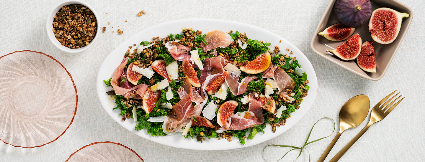 Granola-viikunasalaattia isolla ovaalinmuotoisella tarjoilulautasella. Salaatissa on lehtikaalia, viikunapaloja, parmesaanijuustoa, parmankinkkuviipaleita ja granolaa. K-Ruoka salaattiresepti.