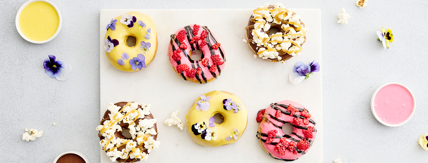 Kuusi värikästä donitsia alustalla, joka on koristeltu orvokeilla.