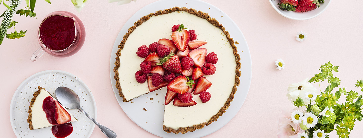 Pannacottapiirakka pyöreällä valkoisella lautasella. Piirakan vaalean pannacottatäytteen päällä on tuoreita mansikoita ja vadelmia. Piirakasta on otettu pala, joka on piirakan vieressä olevalla pienemmällä lautasella. K-Ruoka piirakkaresepti.