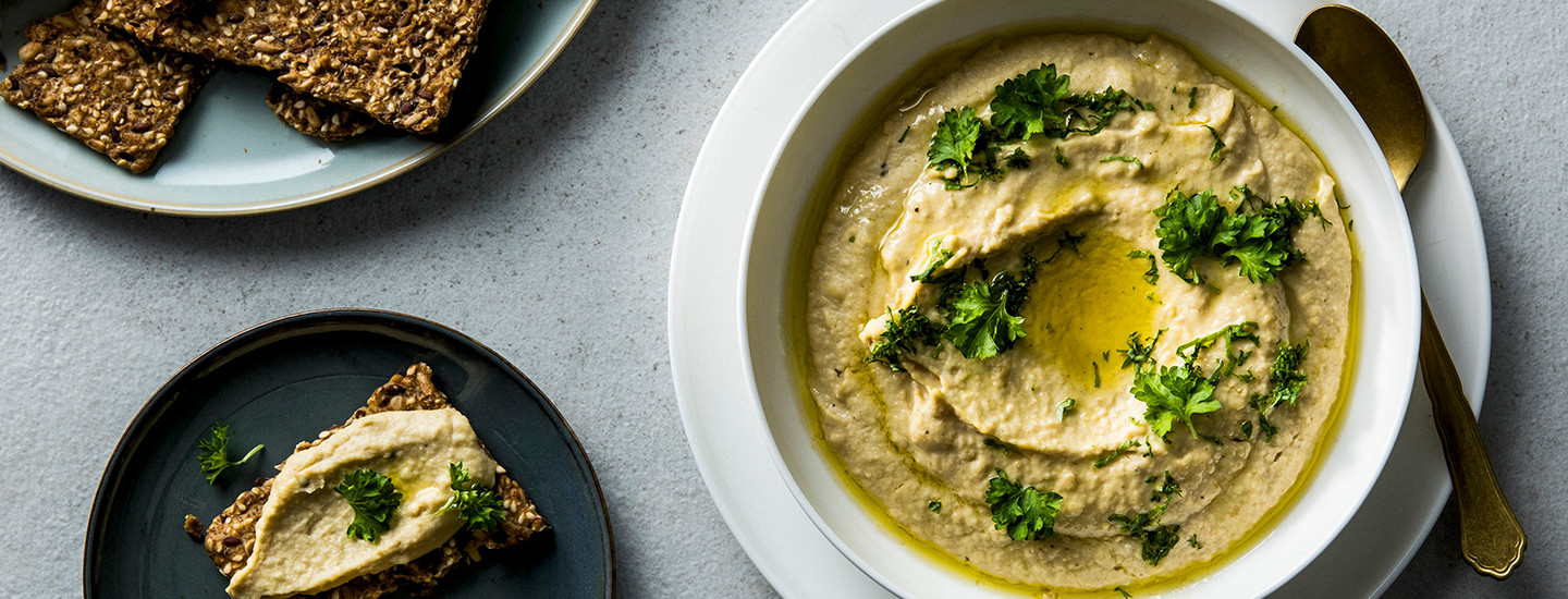 Hummusta valkoisella tarjoilulautasella. Hummuksen päälle on lisätty persiljaa ja oliiviöljyä. Lautasen vasemmalla puolella on mustalla leipälautasella siemenleipää, jonka päällä on hummusta. K-Ruoka hummusresepti.