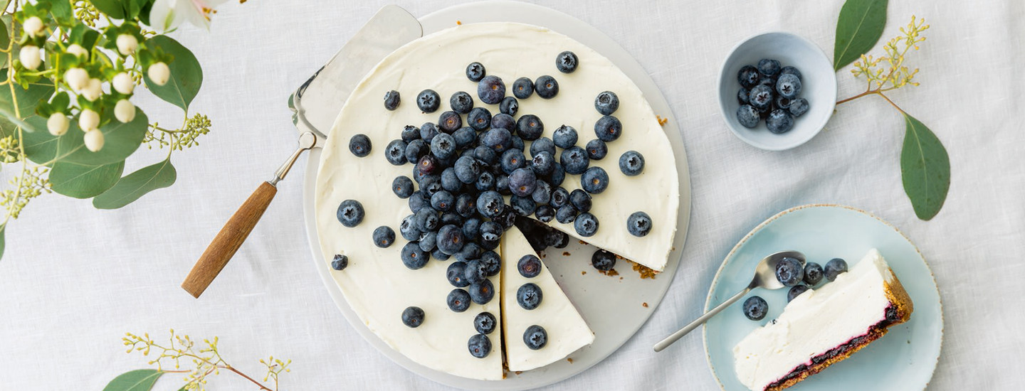 Mustikkahillo-juustokakku valkoisella pyörellä lautasella. Valkoisen juustokakun päälle on lisätty tuoreita mustikoita. Juustokakusta on leikattu palanen, joka on pienellä sinisellä lautasella kakun vieressä. K-Ruoka juustokakkuresepti.