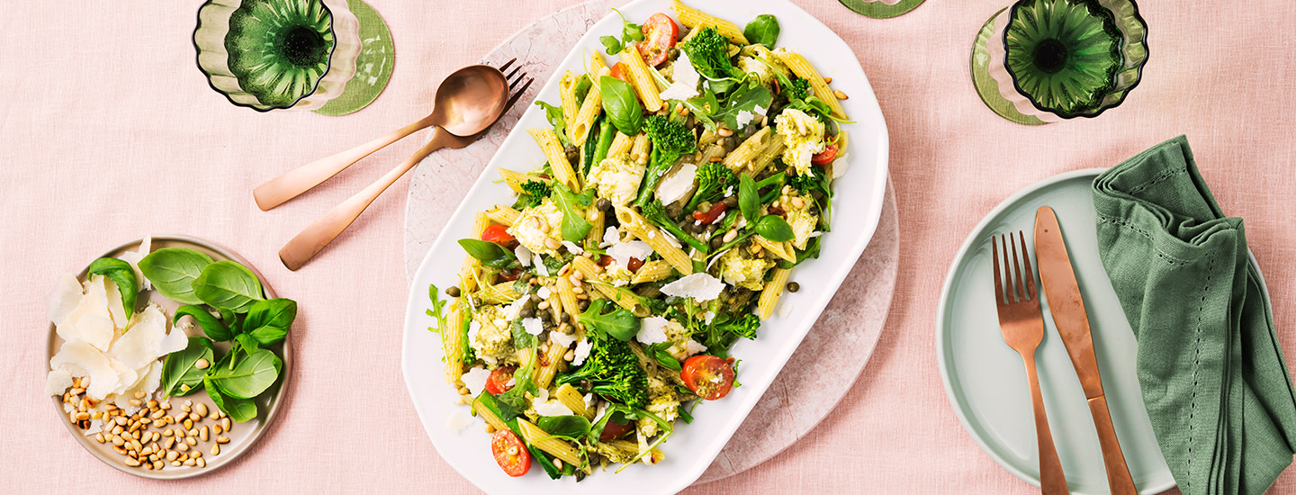 Pestopastasalaattia tarjoilulautasella. Salaatissa on pennepastaa, varsiparsakaalia, mozzarellapaloja, kirsikkatomaatteja, kapriksia, rucolaa ja basilikanlehtiä. K-Ruoka salaattiresepti.