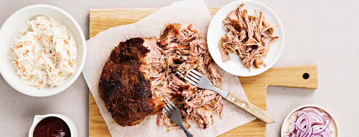 Ylikypsää possua eli pulled porkia puisen leikkuulaudan päällä. Possua on revitty pienemmiksi paloiksi haarukoilla. K-Ruoka resepti.