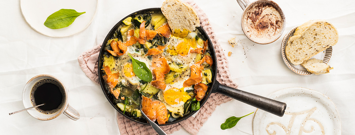 Kylmäsavulohi-munapannu vaaleanpunaisen liinan päällä. Pannun viereen on katettu lämmintä juotavaa kuten kahvia. Myös leipää on katettu pöydälle.