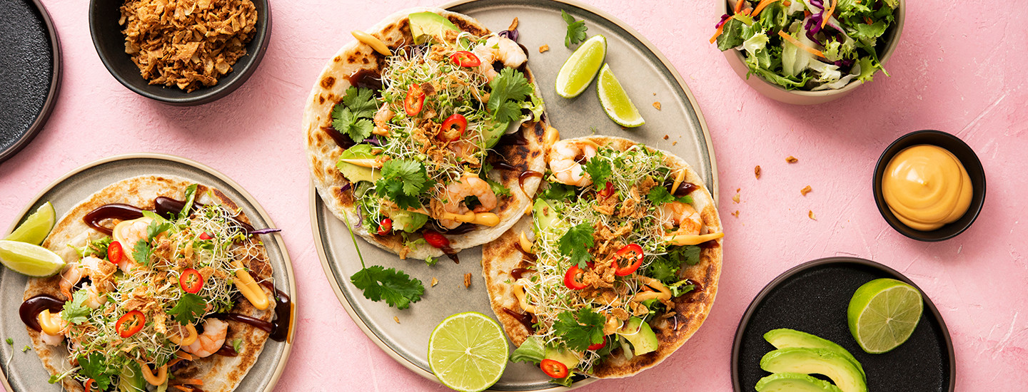 Kaksi shanghai-tavoa isolla pyöreällä lautasella. Kolmas shanghai-taco on vasemmalla toisella pienemmällä lautasella. Tacojen päällä on katkarapuja, avokadoa, kastikkeita ja silmusalaattia. K-Ruoka resepti.