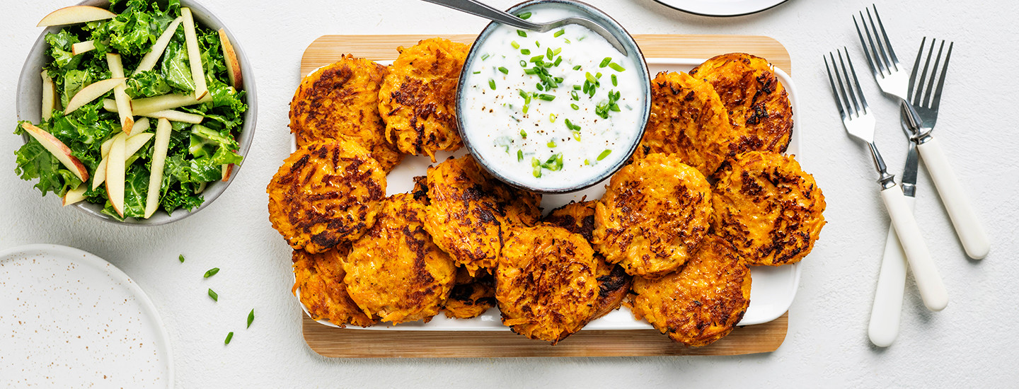 Porkkanapihvejä valkoisen tarjottimen päällä ja tarjotin on puisen leikkuulaudan päällä. Porkkanapihvien keskellä pienessä kulhossa on raejuusto-jogurttikastiketta. K-Ruoka arkiruokaresepti.