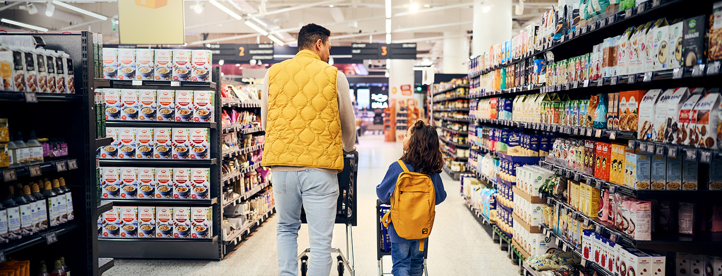 Oletettu isä ja tytär kävelevät K-ruokakaupan leivonta-osastolla. Isällä on keltainen liivi ja tyttärellä keltainen reppu. 