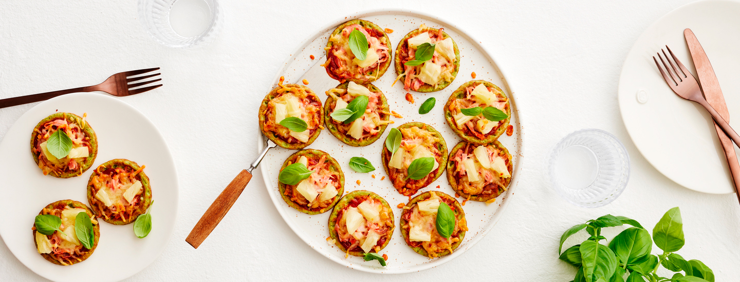 Pinaattilettuja pizzatyylisesti valkoisella lautasella. Pinaattilettujen päälle on lisätty pizzatäytteitä: kinkkua, ananasta, juustoa ja basilikaa. K-Ruoka arkiruokaresepti.