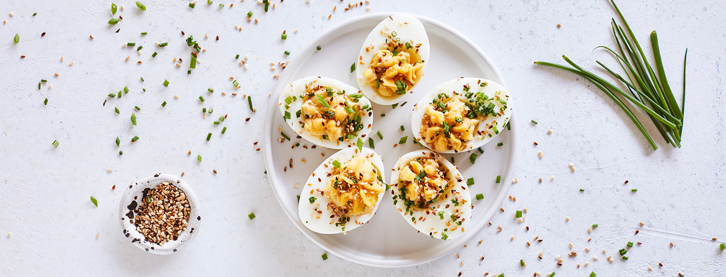 Täytettyjä kananmunanpuolikkaita pienellä valkoisella tarjoilulautasella. Kananmunien päälle on ripoteltu mausteita ja ruohosipulia. K-Ruoka alkupalaresepti.
