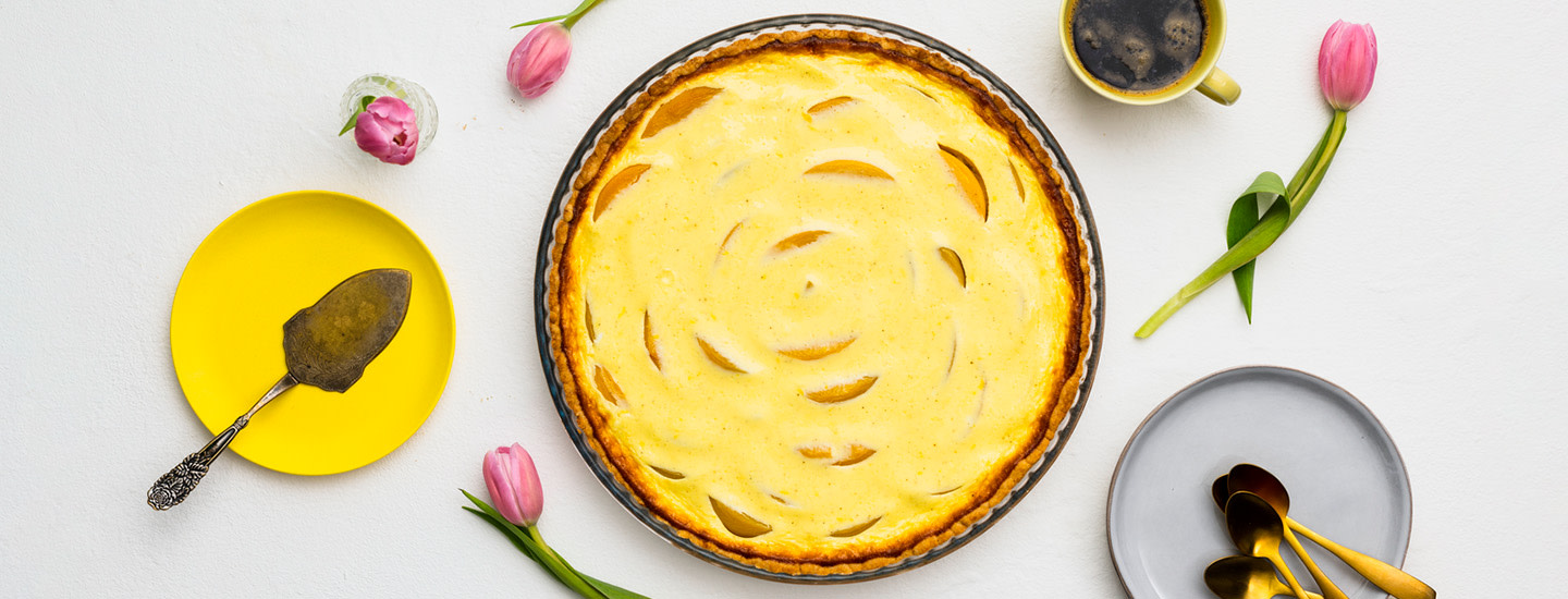 Persikka-rahkapiirakka pyöreässä piirakkavuoassa. Vasemmalla puolella piirakkavuokaa on keltainen pieni lautanen, jossa on kakkulapio. Oikealla puolella piirakkavuokaa on vaaleanpunainen tulppaani pöydällä. K-Ruoka piirakkaresepti.